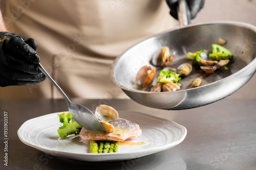 Quadro su tela A chef in black gloves uses a spoon to plate cooked clams over a piece of salmon with asparagus and broccoli