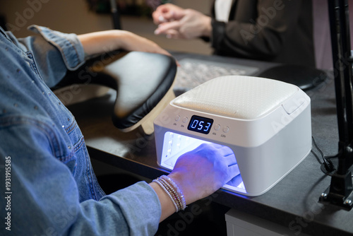 Close-up of female hand in UV nail lamp during gel manicure at beauty salon. Professional nail care, drying gel polish, manicure procedure, self-care and beauty treatment concept.
