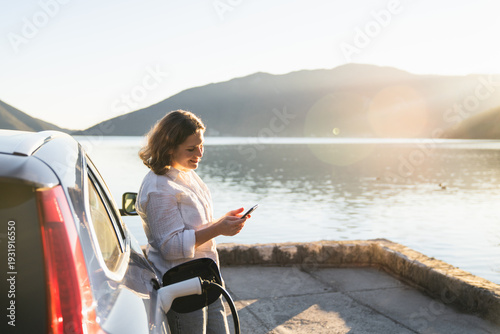 Wallpaper Mural Woman with smartphone next to charging electric car on the seacoast during summer vacation Torontodigital.ca