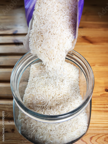 Closeup of dry, uncooked, white basmati rice being poured from a packet into a glass storage jar.