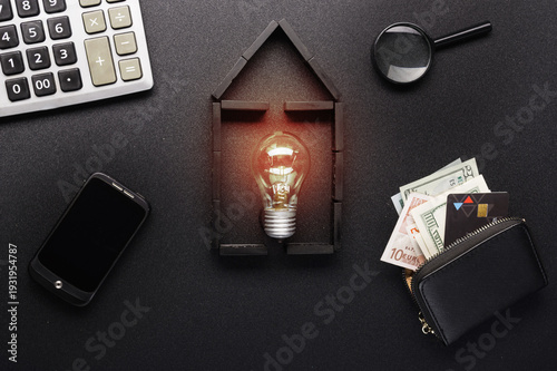 Flat lay domino house with glowing light bulb, money, and calculator on black background.