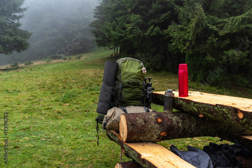 Wooden table from a log house in the forest. A tourist backpack and a thermos on the table. Tourist backpack and thermos on wooden table in forest log house.