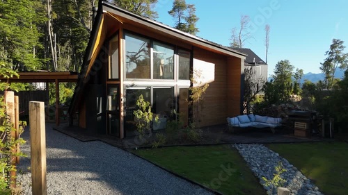 Aerial drone view of a modern luxury cabin with contemporary A-frame architecture, nestled in a lush forest with mountain views and a landscaped garden.