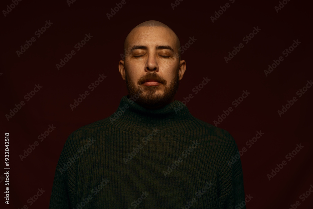 Obraz premium Close-up portrait of a calm bald man with a beard and closed eyes, wearing a dark green turtleneck sweater, posing against a deep burgundy background with dramatic, moody studio lighting.