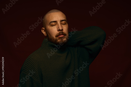 Obraz na plátně Bald man with beard wearing a dark olive green turtleneck sweater, eyes closed in a calm, contemplative pose against a moody dark red background, studio portrait with dramatic low key lighting