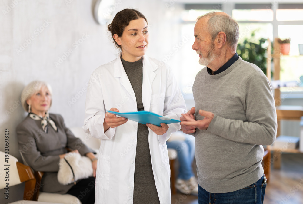 Obraz premium Positive polite young nurse or doctor talking to aged man patient in reception area in background queue of people