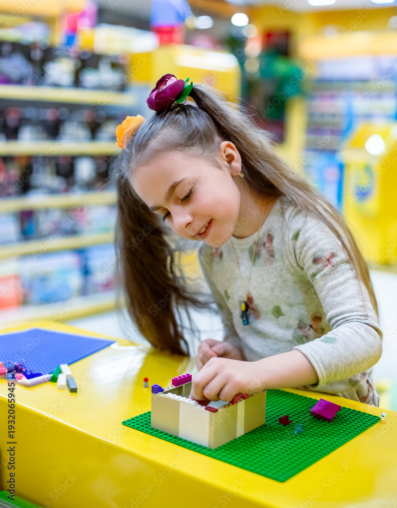 Fototapeta premium Girl concentrating while assembling colorful building bricks green baseplate yellow table toy shop