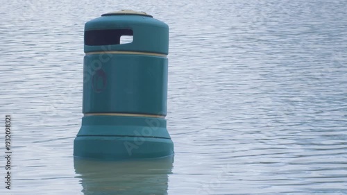 rubbish bin aka trash can surrounded by flood waters, poking up from surface in city park, mute swan swims past in background