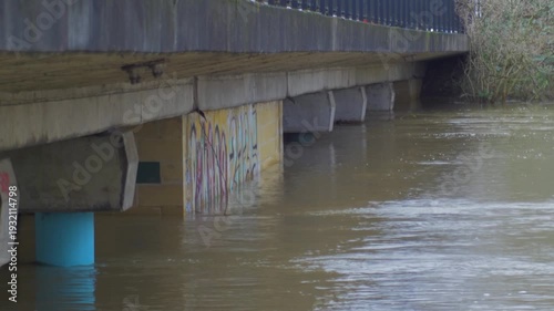 high level of flood water passing through concrete supports of road bridge, riverside city park