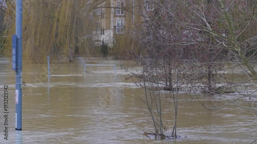 Fast flowing flooded river, muddy brown water, willow trees and residential apartments in background, submerged railing, signpost, lamppost and trees in foreground