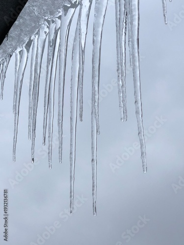 Detailed macro shot of transparent frozen icicles hanging from a roof edge. Soft winter light passes through the ice, creating beautiful light flares and a blurred bokeh background.
