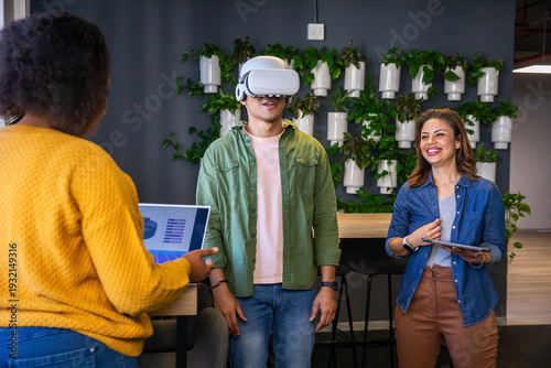 Diverse coworkers in mustard sweater testing VR headset and tablet, pointing to laptop at office