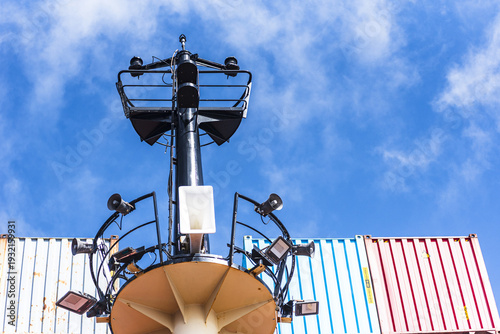 Close-up of a cargo vessel’s foremast with visible navigation lights and technical equipment, stacked containers below and clear blue sky above.