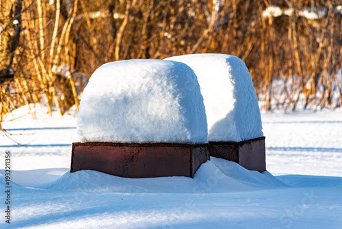 Two heavy-set refuse bins, completely blanketed in pristine snow, stand under a crisp winter sun. Warmly lit distant trees offer a natural contrast