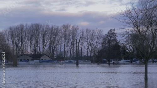 flooded riverside park in later winter light, trees, boats and buildings in background, cloudy stormy sky