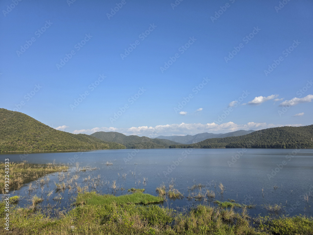Fototapeta premium Serene landscape view of calm lake and green mountain range under clear blue sky. beautiful and peaceful nature background of water and forest on sunny day