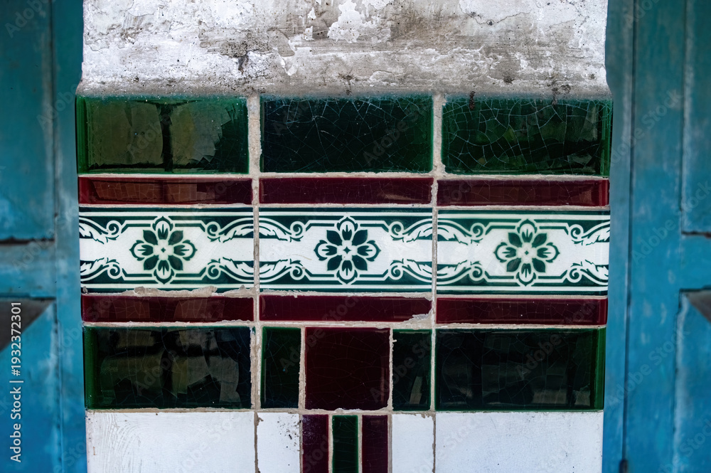 Obraz premium An aged wall column features vibrant green and maroon patterned tiles, framed by a peeling, distressed wall and blue elements, blending decay with color