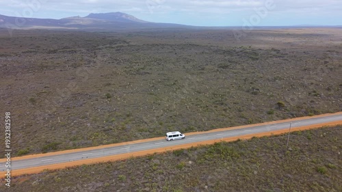 Aerial drone view of an RV motorhome driving along a lonely highway in a semi-desert landscape with distant hills.