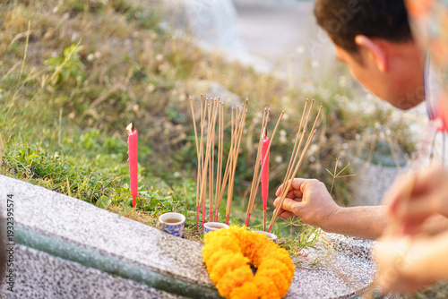 Fotografía Close-up of a man's hand placing burning incense sticks at a Chinese grave during Qingming Festival, honoring ancestors with red candles, tea offerings, and marigold garlands in Thailand