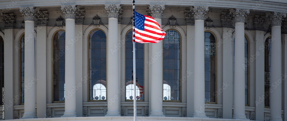 Naklejka premium Capitol with american flag near dome landmark. American flag near capitol with classical columns. Us flag on capitol as political symbol. Capitol with american flag for national background.