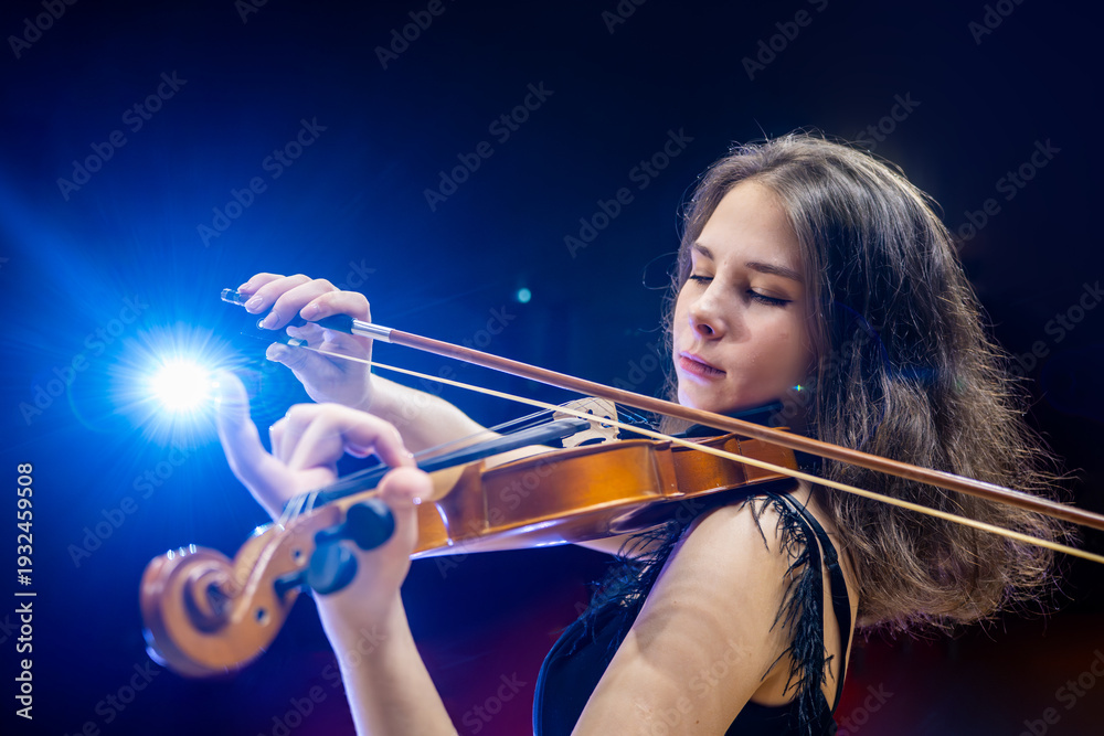 Fototapeta premium young woman in a dress plays the violin on stage under spotlight beams, an elegant and professional performance.