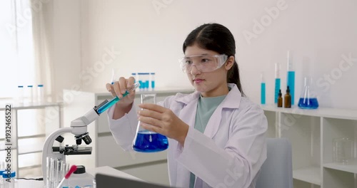 Asian primary student in labcoat using glass rod to stir chemical liquid in beaker during school science experiment, focusing carefully while sitting beside microscope and testtubes in laboratory