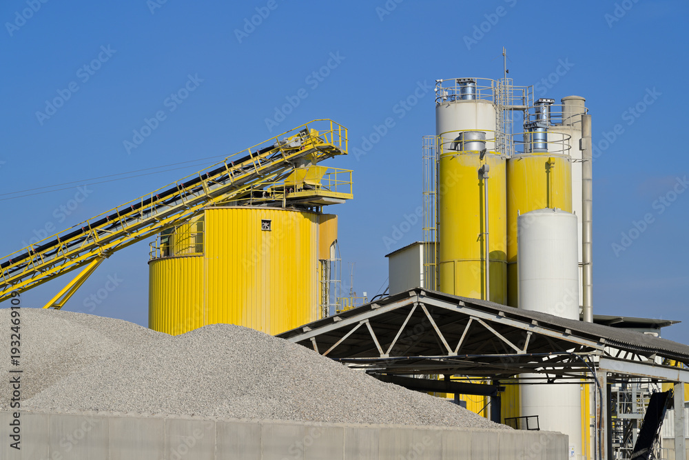 Obraz premium Industrial facility processing aggregates with conveyor belt and silos. Large industrial complex featuring silos, conveyor belts, and piles of raw material under a blue sky.