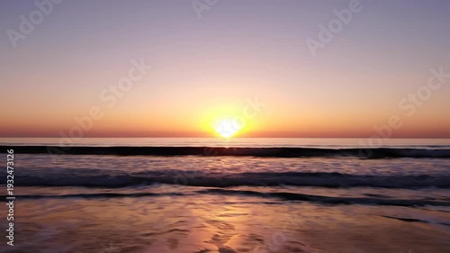 Sunrise over calm ocean waves, showcasing vibrant colors reflecting on water surface, with sandy beach in foreground and clear sky above