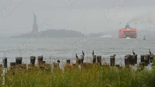 Statue of Liberty on Liberty Island from Brooklyn, New York City, United States. Ferry to Staten Island and birds on old wooden pier piles remains. Rainy foggy day weather. Orange boat water transport