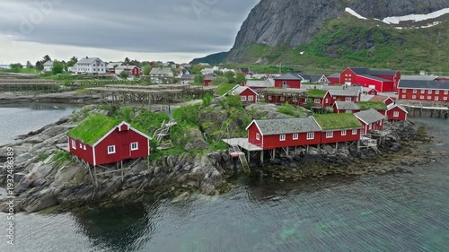 Aerial tracking shot over Hamnøy fishing village in Lofoten Islands, Norway. Norwegian red cabins and coastline framed by steep mountains and calm northern waters.