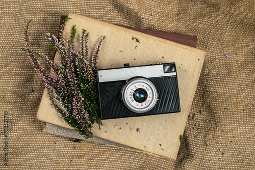 Photography Top view of a vintage analog camera resting on stacked old books with pink heather flowers on textured burlap fabric