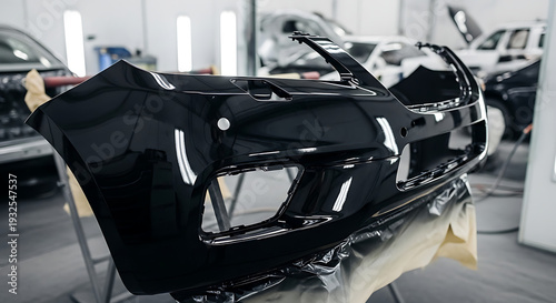 A black car bumper being repaired or manufactured in a well-lit automotive workshop with various cars in the background