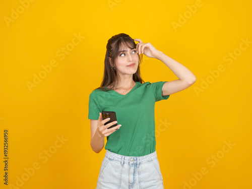 Thoughtful young woman in green t-shirt holding smartphone and scratching her head while looking away. Concept of forgetting a password, choosing an app, or searching for information against yellow ba