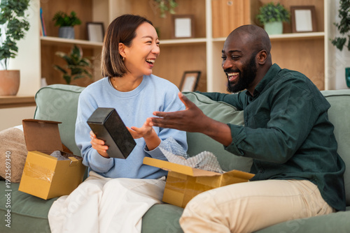 A multiracial couple relaxes on the couch, reviewing an online order as they unpack a freshly delivered box of perfume.
