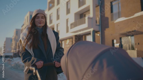 Caucasian woman pushing baby stroller. Wearing knitted hat and warm coat. Looking at child while walking slowly at winter street. Modern residential district. Caring mother enjoying walk outdoors.