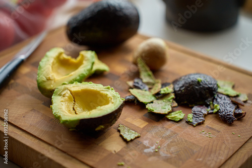 Avocado halves with pit, peel scraps and knife on wooden cutting board. Concept of food preparation, healthy cooking and guacamole