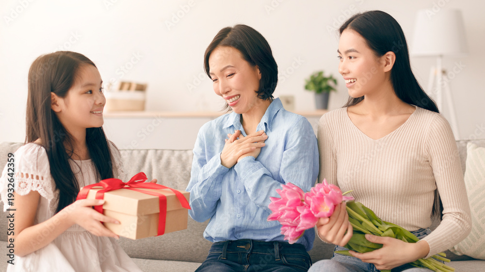 Fototapeta premium Two children surprise their mother with a gift and flowers. They sit on a sofa in a cozy living room. The mother looks happy and surprised. It is a joyful moment of love and appreciation.
