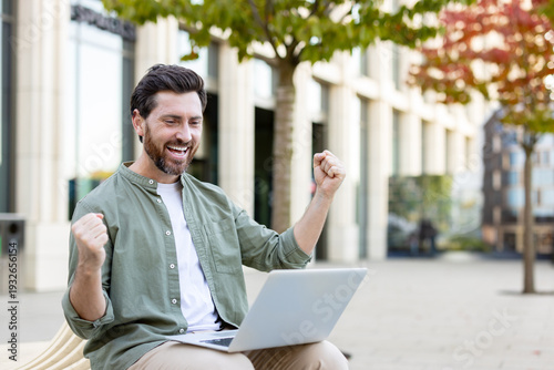 Εκτύπωση καμβά Excited man cheering success while looking at his laptop, sitting on a bench in
