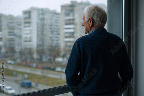 senior man standing by large window looking outside, photographed from behind, tense shoulders and hands in pockets, rainy city background, soft diffused daylight, clean apartment interior, commercial