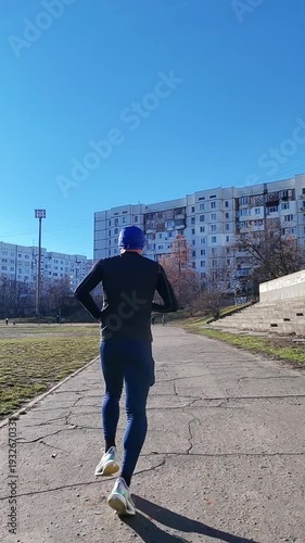 Athletic young man running on the road.Dressed in blue pants, black jacket, blue hat. marathon training. Back view.Healthy, active athlete or speed runner for cardio performance
