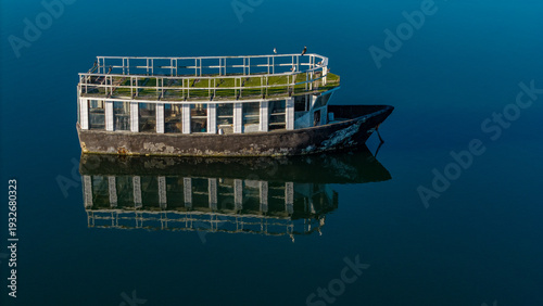 Aerial view of a weathered, two-story boat with a white-railed upper deck, sits still reflecting in the dark, glassy water., Knysna, Western Cape, South Africa.