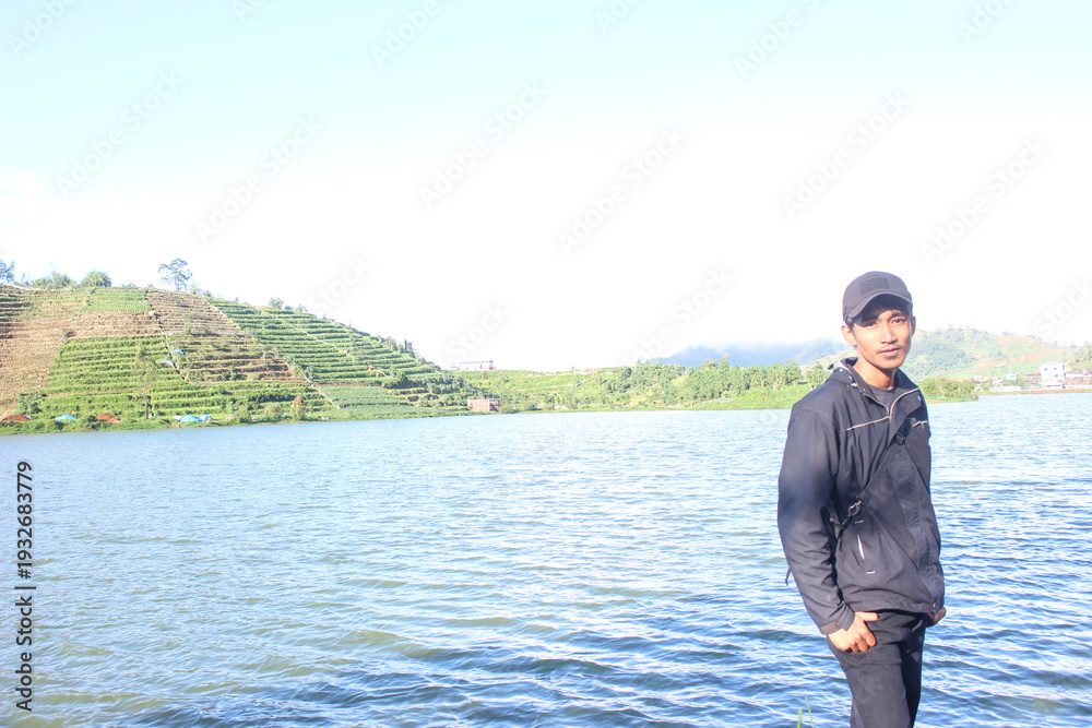 Fototapeta premium Young man standing by large lake near terraced hillside agriculture landscape