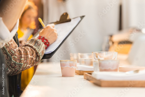 Obraz na plátně A professional judge or barista taking notes on a clipboard during a coffee cupping or sensory evaluation at a cafe competition, focusing on artisanal ceramic cups and a warm, blurred background