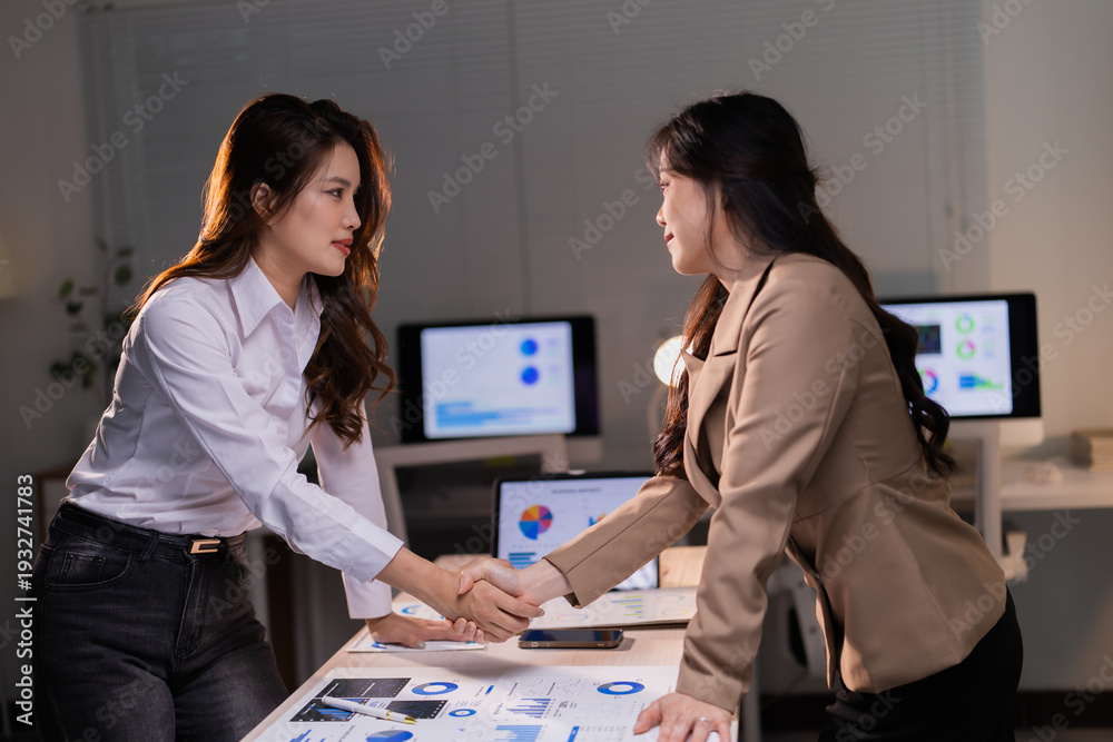 Fototapeta premium Two professional businesswomen are shaking hands over financial documents and digital screens displaying charts in an office, symbolizing a successful partnership, agreement, and collaboration