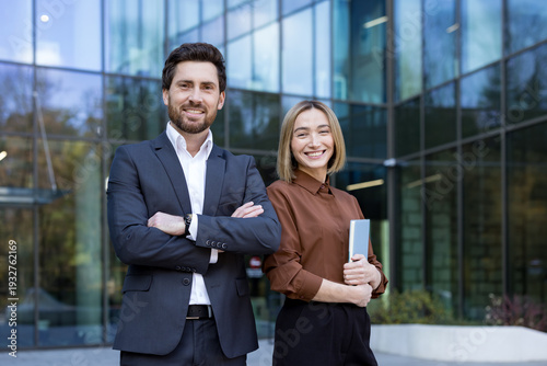 Fotografi Confident business man and woman standing by modern office building, representin