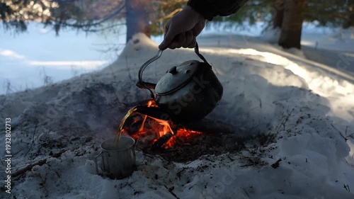 Hot tea from a camping kettle is poured into a metal mug standing on the snow. Winter camping. Hot tea on a winter hike. Bushcraft and wilderness survival concept.