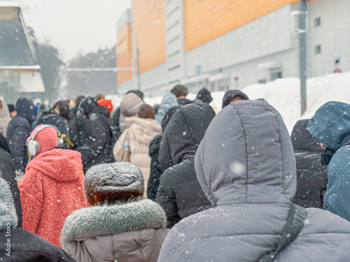 People standing in queue during snowfall near urban building, wearing winter jackets and hoods, backs turned to camera in cold weather. Concept of queue, waiting line, social issue, crowd
