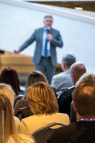 Blurred speaker presents to focused crowd during corporate event in sleek conference space. Audience seated closely listens as ideas, strategies, insights unfold on stage