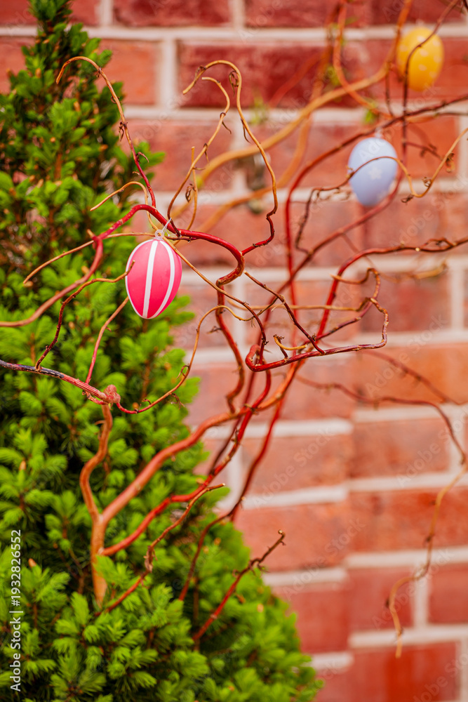 Fototapeta premium Brightly colored Easter eggs dangle from the whimsical branches against a rustic brick wall