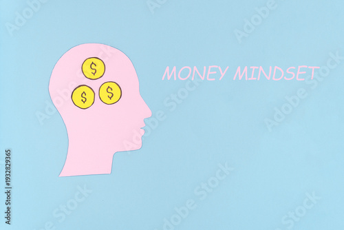 A graphic illustration of a human head silhouette in pink with three dollar signs inside, symbolizing a money mindset concept against a light blue background.